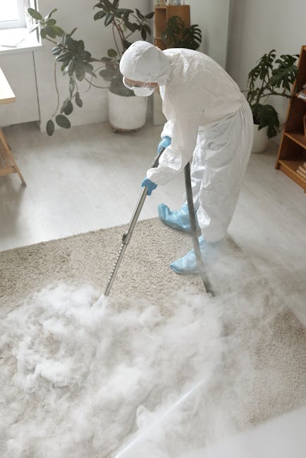 A professional cleaner dressed in white protective coveralls, gloves, and a face mask uses a vacuum or steam cleaning machine to deep clean a plush, light-colored carpet in a well-lit living room. The