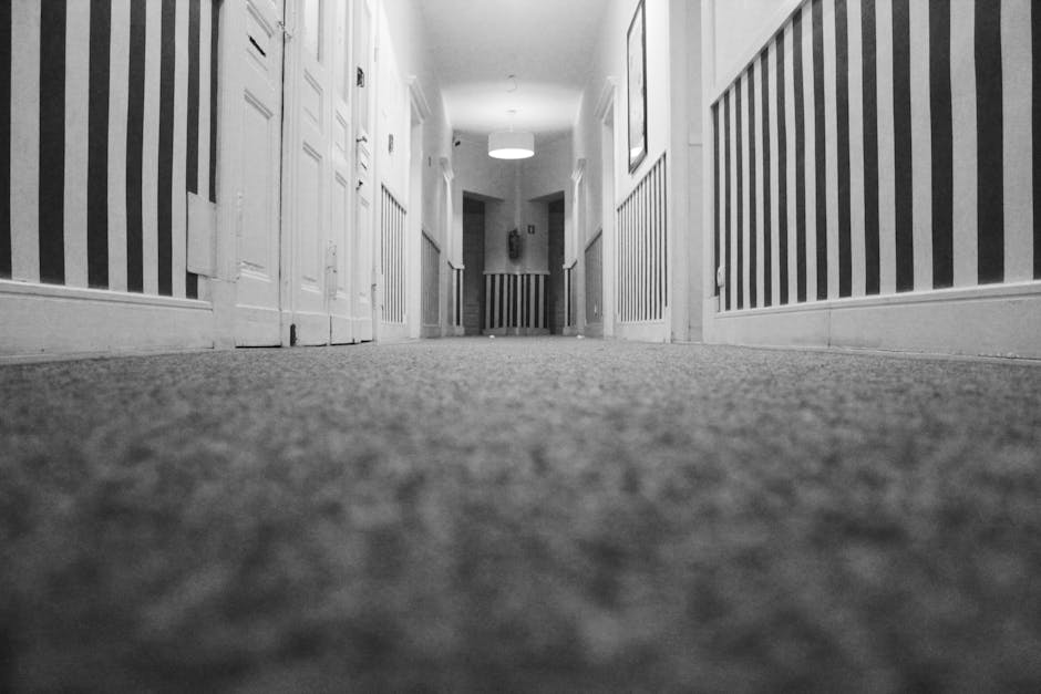 A low-angle view of a clean, well-maintained corridor in a residential property, featuring a plush grey carpet that appears free of dust and stains, extending through the length of the hallway. The corridor is lined with white paneled doors and decorative striped wallpaper on the walls, creating a neat and hygienic appearance. Soft ambient lighting from ceiling fixtures illuminates the space evenly, highlighting the smooth texture of the carpet. Visible at the end of the corridor is a small, dark area that suggests the continuation of the house layout. This image emphasizes top-quality surface cleaning and deep cleaning processes, which are part of professional domestic cleaning services provided by [COMPANY_NAME], ensuring thorough sanitisation and maintenance of interior carpets and flooring surfaces.