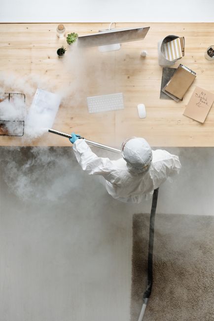A professional cleaner dressed in white protective coveralls, gloves, and a hood is performing deep cleaning or sanitisation of a carpeted office area using a steam or misting tool. The image is taken from an overhead perspective, showing a wooden desk or counter with a computer monitor, keyboard, mouse, and various office supplies including notebooks and a small potted plant. The cleaner is standing on a light grey carpet, with the process creating visible mist or steam over the carpet surface, indicating thorough surface cleaning and sanitation. The workspace appears organized and clean, highlighting the importance of hygiene maintenance in commercial environments, with [COMPANY_NAME] providing specialist cleaning services. The scene emphasizes the importance of meticulous surface cleaning to maintain a hygienic office space, aligning with the page topic on commercial office cleaning rates in Kingston upon Thames.
