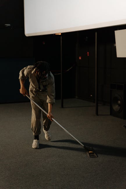 A professional cleaner dressed in white protective coveralls, gloves, and a hood is performing deep cleaning or sanitisation of a carpeted office area using a steam or misting tool. The image is taken from an overhead perspective, showing a wooden desk or counter with a computer monitor, keyboard, mouse, and various office supplies including notebooks and a small potted plant. The cleaner is standing on a light grey carpet, with the process creating visible mist or steam over the carpet surface, indicating thorough surface cleaning and sanitation. The workspace appears organized and clean, highlighting the importance of hygiene maintenance in commercial environments, with [COMPANY_NAME] providing specialist cleaning services. The scene emphasizes the importance of meticulous surface cleaning to maintain a hygienic office space, aligning with the page topic on commercial office cleaning rates in Kingston upon Thames.