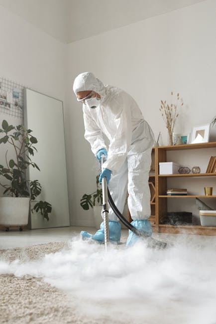 A professional cleaner dressed in full white protective suit, mask, and gloves is performing a deep cleaning process on a beige carpet in a modern, well-lit living room. The cleaner is using a steam or fogging device that emits a white vapor or fog over the carpet surface. The room features a light-colored wall, a large mirror, and a wooden bookshelf with decorative items, plants, and books. Natural daylight illuminates the space, highlighting the pristine condition of the carpet. This scene illustrates surface cleaning and sanitisation services typically offered by [COMPANY_NAME], emphasizing hygiene and thorough domestic cleaning in a residential setting.