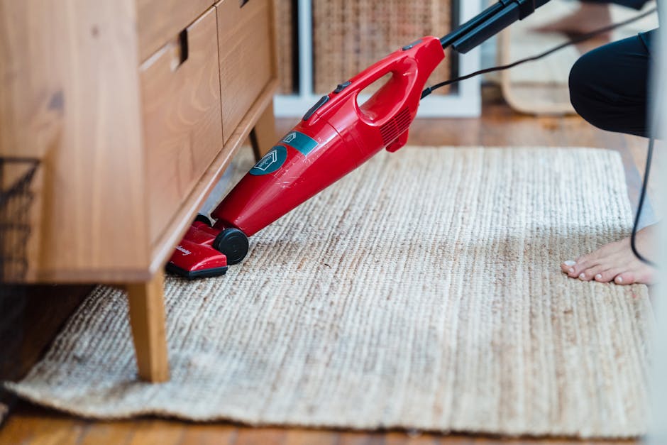 A woman with dark hair tied back and wearing glasses is performing surface cleaning in a domestic setting, using a handheld vacuum cleaner to vacuum a gray fabric cushion placed on a sofa. She is dres