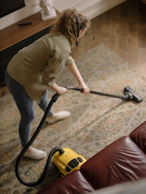A woman vacuuming a patterned area rug in a living room during a domestic cleaning session. She is wearing a green jacket and dark jeans, holding a handheld vacuum cleaner with a yellow body and black