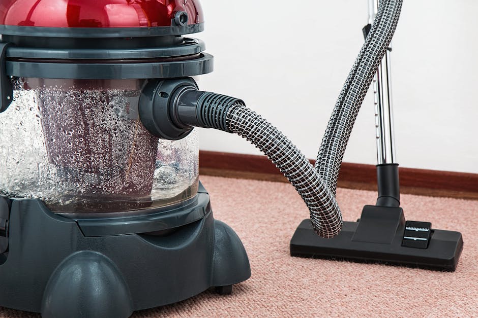 A close-up of a vacuum cleaner with a transparent dust container filled with debris, positioned on a beige carpet in a residential room. The vacuum's hose and long extension wand are visible, with the hose connected to the machine. The carpet appears clean and plush, and the background features a plain white wall with a wooden skirting board. The scene emphasizes surface cleaning and maintenance, illustrating the use of professional equipment for deep cleaning and sanitisation, as might be provided by [COMPANY_NAME] for upholstery and carpet hygiene. The lighting is soft and even, highlighting the cleanliness of the area.
