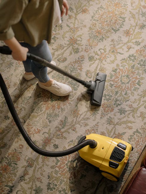 A person using a vacuum cleaner with a long metal wand attachment on a traditional floral-patterned area rug in a room with timber flooring and natural lighting. The rug features pastel shades of beige, green, and pink, and appears clean and well-maintained. The individual is dressed casually in jeans, white shoes, and a beige jacket, operating the vacuum to perform deep cleaning or surface cleaning of the carpet. The yellow vacuum cleaner is positioned on the floor nearby. This scene illustrates domestic rug cleaning as part of general surface or upholstery sanitisation, emphasizing professional cleaning practices by [COMPANY_NAME].